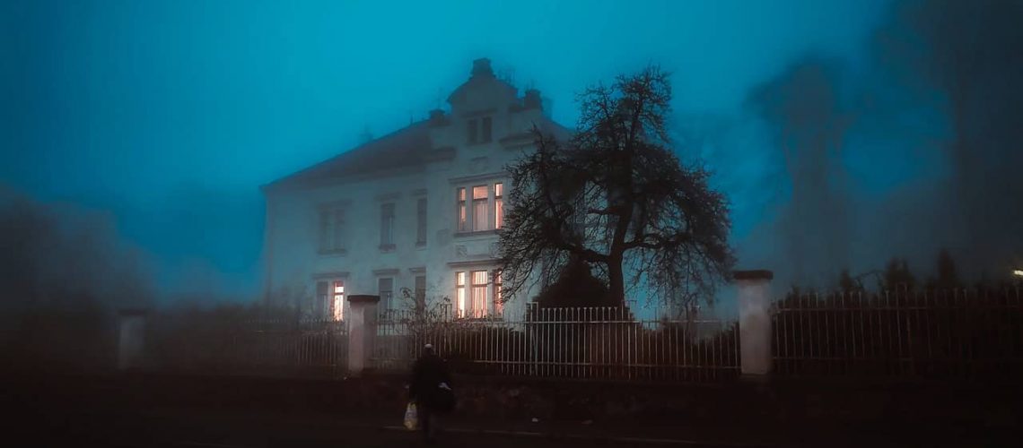Haunted house shrouded in fog with eerie lighting and dark trees, creating a spooky atmosphere.