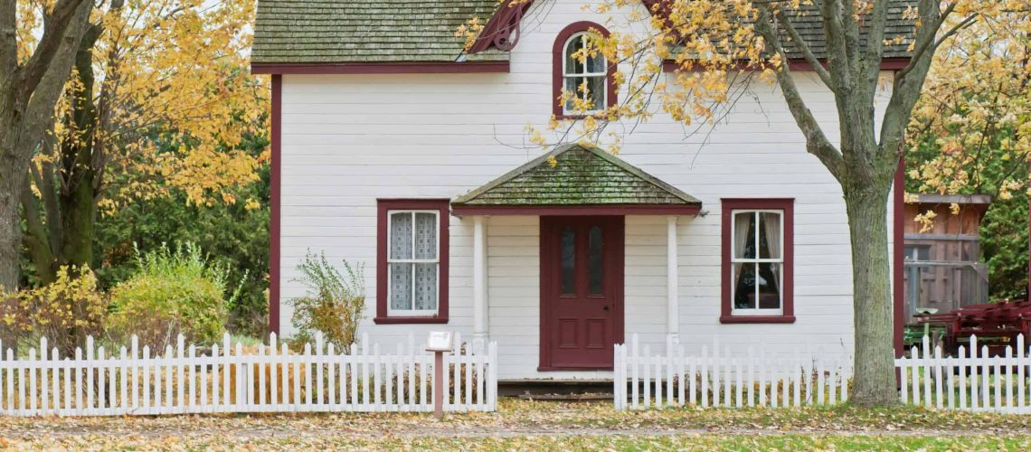Beautiful white house with a red door surrounded by autumn trees and a white picket fence.